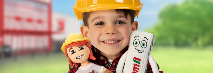 Kind mit Bauhelm hält zwei Plüschfiguren, nämlich Tom und Zolli in der Hand.